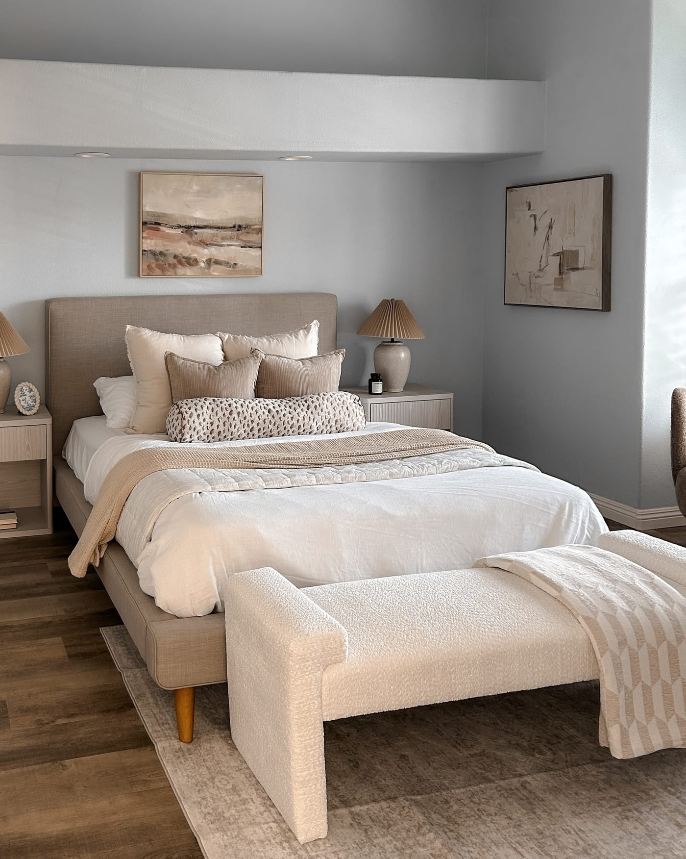 Staged primary bedroom with an upholstered bed, boucle bench, and coastal art