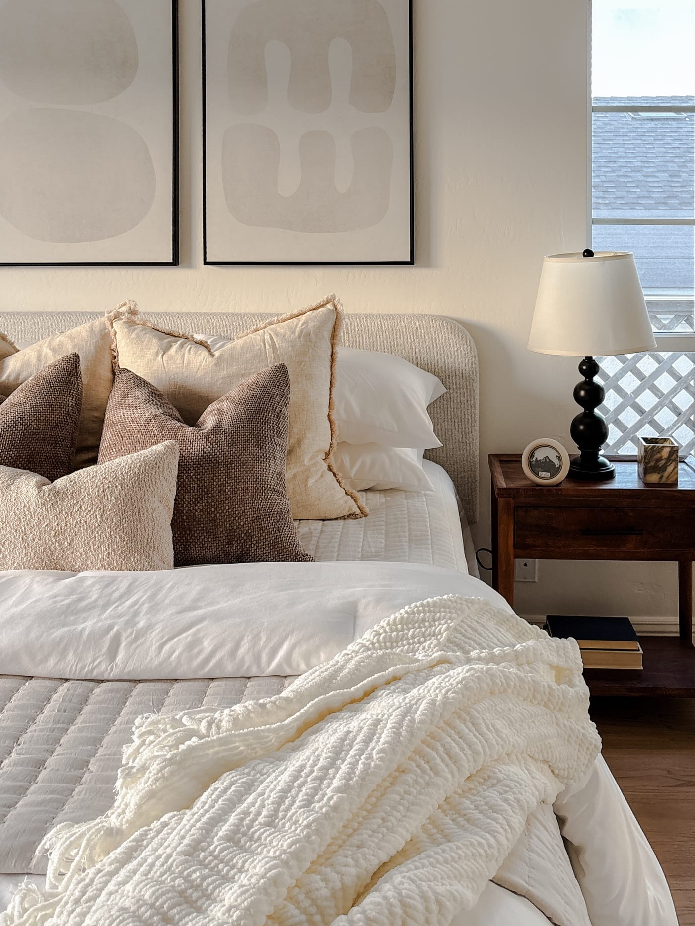 Staged bedroom with an upholstered headboard, layered neutral pillows, and an abstract diptych